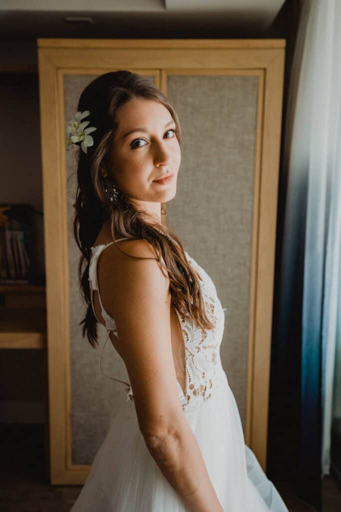 A woman in a white dress with lace details and a flower in her hair stands by a window, looking over her shoulder.
