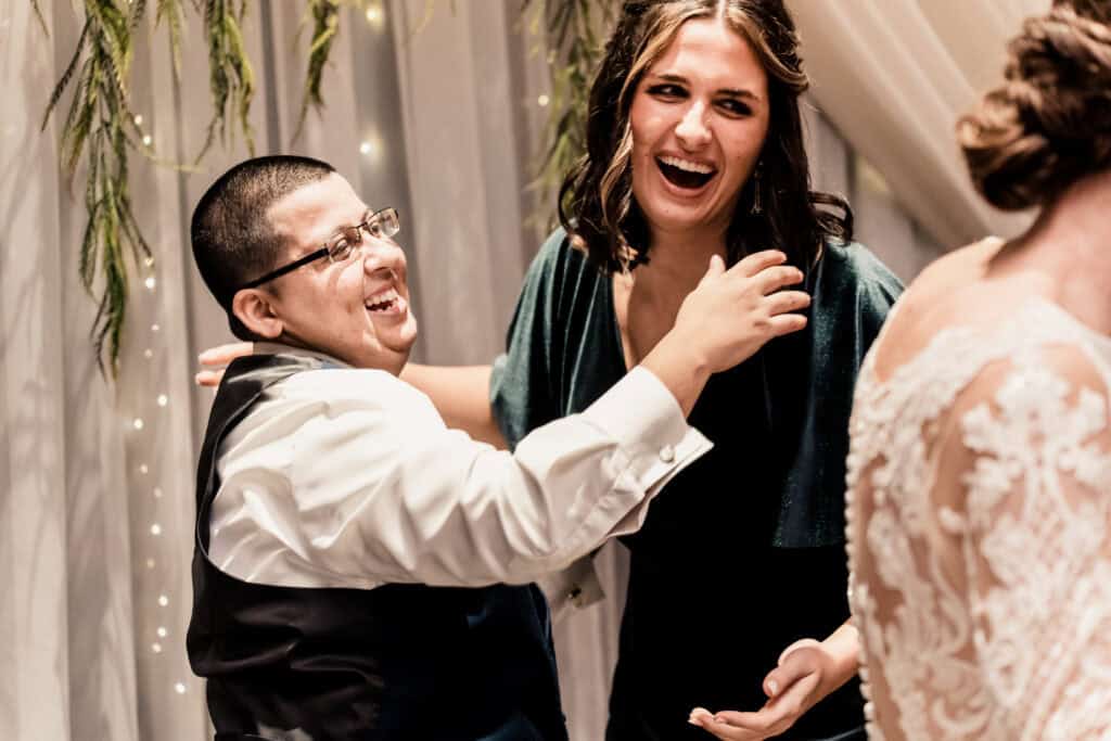 Two people, dressed formally, share a joyous moment and laugh together at an event. A third person in decorative attire is partially visible in the foreground.