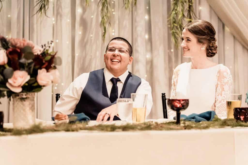A man and a woman sitting side by side at a decorated table, smiling and dressed in formal attire, with a floral arrangement in the foreground.
