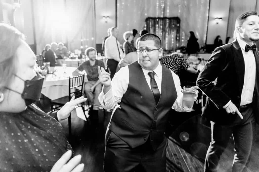 A person in a vest and tie dances while holding a drink, surrounded by others at an indoor event with lights and decorations.