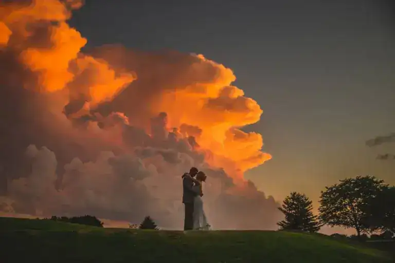 A couple embraces on a grassy hill at sunset, with dramatic orange and pink clouds filling the sky behind them. Trees are silhouetted in the background, perfectly capturing a moment that any La Crosse WI Wedding Photographer would love to frame. In La Crosse, WI.
