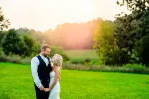 A couple stands together in a grassy field at sunset, holding hands and looking at each other. The man is wearing a vest and tie while the woman is in a white dress. Trees and fields are in the background, capturing this romantic moment as a perfect Denver Colorado Wedding photographer would.