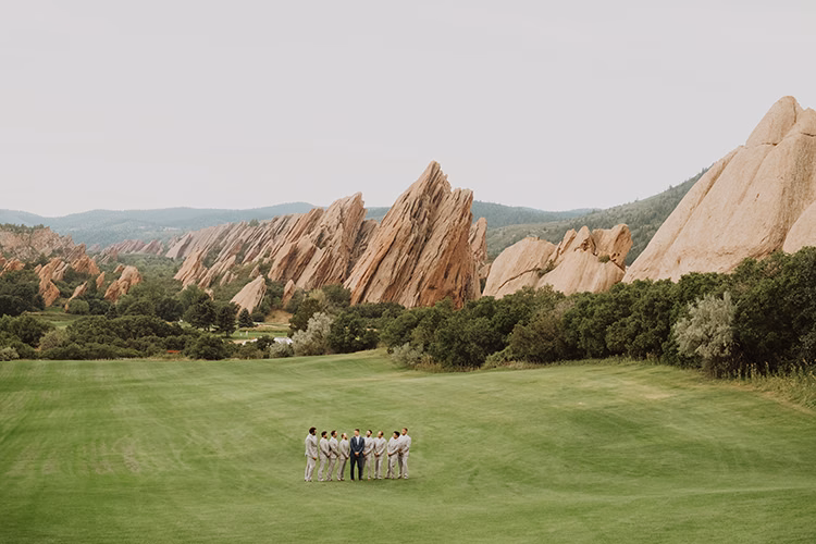 A small wedding party stands on a green lawn at an Arrowhead Golf Club Wedding, with dramatic, jagged red rock formations and hills in the background.