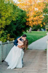 A groom in a navy suit dips and kisses a bride in a white wedding dress on a wooden bridge at the WELLSHIRE EVENT CENTER Wedding Venue, surrounded by green and autumn-colored trees.