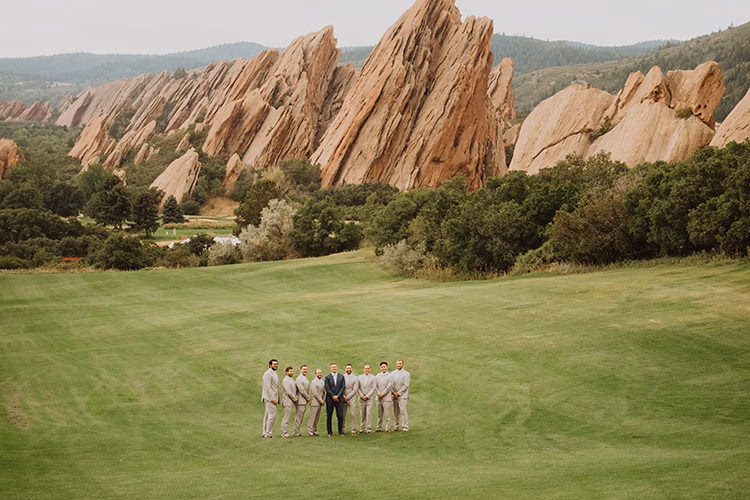 A group of men in suits stand on a green lawn at an Arrowhead Golf Club Wedding, with tall, jagged red rock formations and trees in the background.
