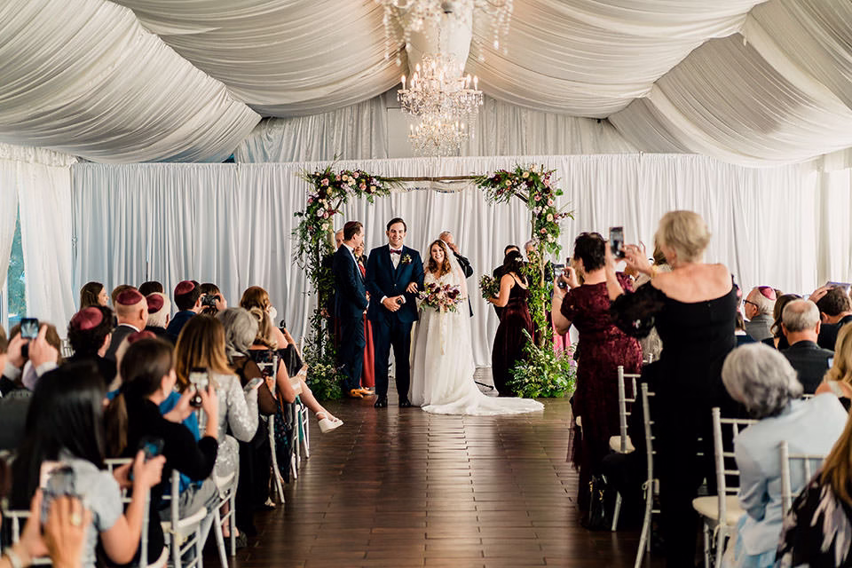 A bride and groom stand under a floral wedding arch at WELLSHIRE EVENT CENTER Wedding Venue, with guests seated and some taking photos inside a beautifully decorated venue with white draped ceilings.