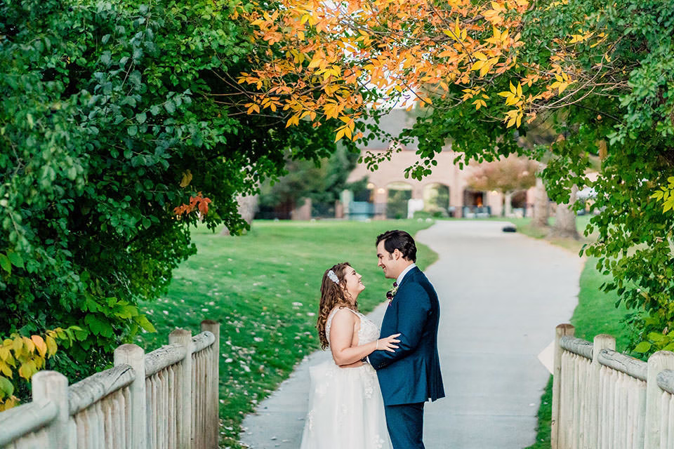 A bride and groom stand facing each other on a wooden bridge at the WELLSHIRE EVENT CENTER Wedding Venue, surrounded by green and autumn-colored leaves, with a path and buildings in the background.