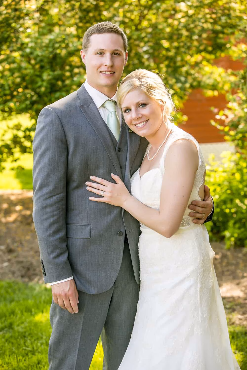 A bride and groom pose together outdoors, both smiling. The groom wears a gray suit and the bride wears a white dress. Greenery and sunlight are visible in the background.