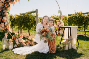 A bride in a white dress and a young girl in a rust-colored dress pose together outdoors, surrounded by flowers and greenery.