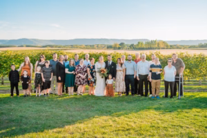A large group of people, including a bride and groom, pose together outdoors in a vineyard with hills in the background on a sunny day.