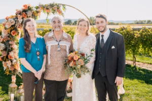 Four people stand outdoors in front of a floral wedding arch; one couple is dressed in wedding attire, while the other two are dressed in more casual clothing.