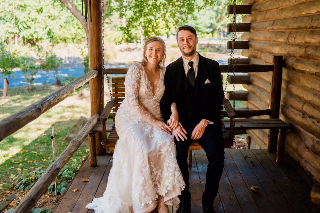 A woman in a white lace dress and a man in a black suit sit side by side on a wooden porch swing attached to a log cabin, surrounded by greenery.