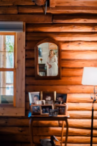 A woman in a white dress is reflected in a wall mirror inside a log cabin; framed photos and a lamp are visible in the foreground.