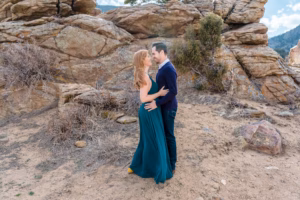 A woman in a green dress and a man in a navy sweater stand facing each other, embracing, in front of rocky terrain outdoors.