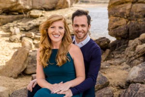 A woman in a green dress and a man in a navy sweater sit together on rocks outdoors, smiling at the camera with a rocky shoreline in the background.