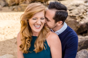 A man embraces a woman from behind while she laughs with her eyes closed. They are outdoors near rocks and sand.