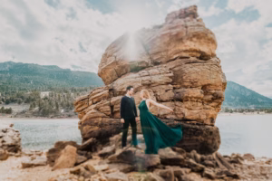 A man and woman stand on rocky terrain near a large rock formation by a lake, with mountains and trees in the background. The woman wears a flowing green dress.