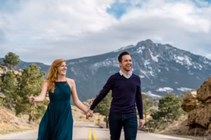 A woman in a green dress and a man in a dark sweater hold hands and walk down a road with mountains and trees in the background.