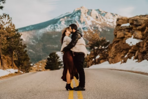 A couple stands and kisses on a mountain road with snow on the ground and rocky cliffs in the background.