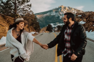 A woman and a man hold hands and walk down a mountain road with snow and rocky cliffs on either side, with a large mountain in the background.
