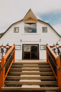A wide staircase with black railings and orange banisters leads up to double glass doors of a white barn-style building with green trim.