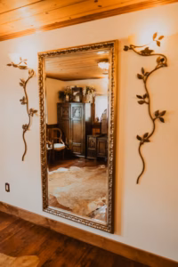 Large ornate mirror on a wall reflects a room with wooden furniture, a chair, a cowhide rug, and a ceiling with wooden paneling. Decorative metal vines flank the mirror.