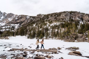 Two people walk on snow near a rocky, forested mountain landscape under a cloudy sky.