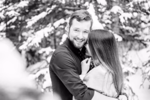 A man and woman embrace outdoors in a snowy forest, both smiling. The image is black and white.
