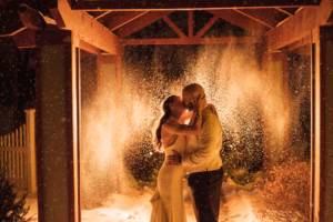 A couple in formal attire embraces and kisses under a wooden structure at night, surrounded by illuminated falling snow—capturing the magic of Colorado Parks Wedding Venues.