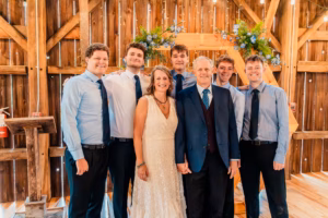 A group of seven people, including a woman in a white dress and a man in a suit, stand together smiling in front of a wooden backdrop decorated with flowers.