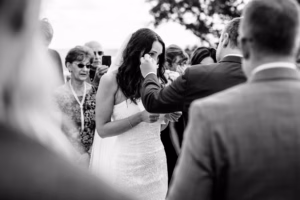 A bride in a strapless gown wipes her eye during an outdoor wedding ceremony, surrounded by guests, in black and white.