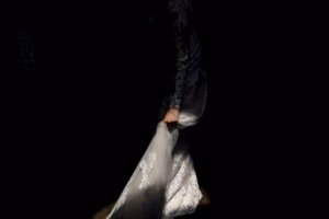 A person in a lace dress is partially illuminated against a dark background, holding the bottom of the dress with one hand.
