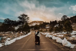 Two people embrace on a paved road surrounded by snow and trees, with mountains and a dramatic sky in the background at sunset.