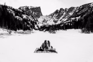 Two people sit on a blanket in the snow, facing each other, with snow-covered mountains and pine trees in the background.