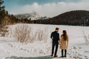 A man and woman hold hands while standing on a snowy path, facing a frozen lake surrounded by mountains and pine trees.