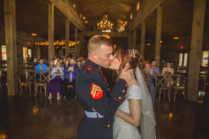 A bride and groom kiss on the dance floor in a rustic venue, with guests seated and watching in the background.