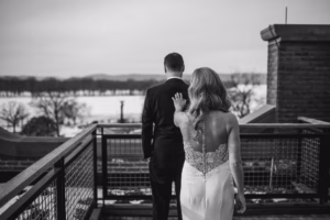 A woman in a wedding dress approaches a man in a suit from behind on an outdoor balcony at Somerby Golf Club, with a snowy landscape in the background, capturing the magic of their winter wedding.