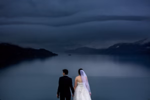 A bride and groom stand hand in hand, facing a calm lake with mountains and dramatic clouds in the background—an idyllic scene found at Colorado Parks wedding venues.