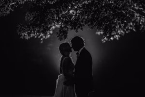 A couple stands under a tree at night, silhouetted and facing each other closely, with soft backlighting highlighting their outlines.