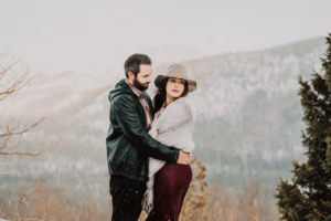 A man and woman stand together outdoors in winter attire, with snow falling and mountains in the background.