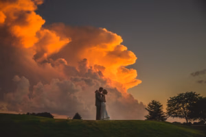 A couple in wedding attire embraces on a grassy hill at sunset, with dramatic orange clouds filling the sky behind them—an unforgettable scene perfect for those seeking Colorado Parks Wedding Venues.