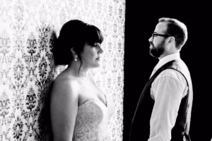 A woman in a strapless dress stands against a patterned wall, facing a bearded man in glasses and a vest. Both look serious. The image is in black and white.
