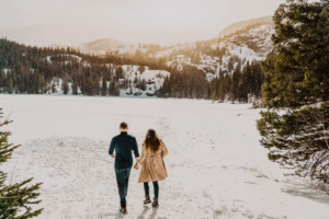 A couple walks hand in hand across a snowy landscape surrounded by trees and mountains under a cloudy sky.
