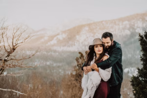 A couple stands outdoors in front of a snowy mountain landscape; the man hugs the woman from behind as she looks at the camera.