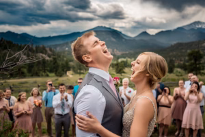 A bride and groom laughing joyfully during an outdoor wedding ceremony, with guests clapping and mountains in the background under a cloudy sky.