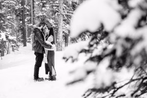 A couple stands close together in a snowy forest, facing each other and holding hands. Snow-covered trees fill the background and foreground.