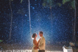 A couple stands on a sandy beach at night, facing each other and illuminated by a bright light as water or rain falls around them—a magical scene reminiscent of Colorado Parks wedding venues.