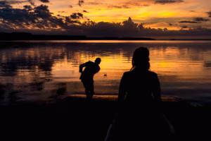 Two people are silhouetted against a sunset over a lake in one of the scenic Colorado Parks Wedding Venues; one skips a stone across the water while the other stands nearby watching.