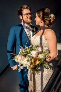 A bride in a lace dress holding a bouquet stands beside a groom in a blue suit and floral tie, both looking at each other and smiling in natural sunlight.