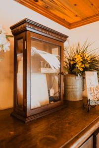 A wooden display case with gloves and a "LOVE" sign inside sits on a wooden surface next to a metal vase with yellow flowers and a pamphlet on a stand.